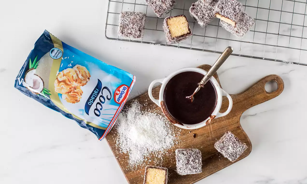 Cubos de bizcocho dorado bañados en chocolate brillante y cubiertos de coco blanco rallado sobre tabla de madera y rejilla metálica