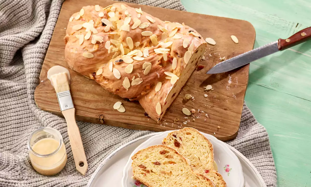 Goldbrauner, geflochtener Striezel mit Mandelsplittern auf Holzbrett, daneben Messer, Glasur mit Pinsel und zwei Scheiben auf Teller