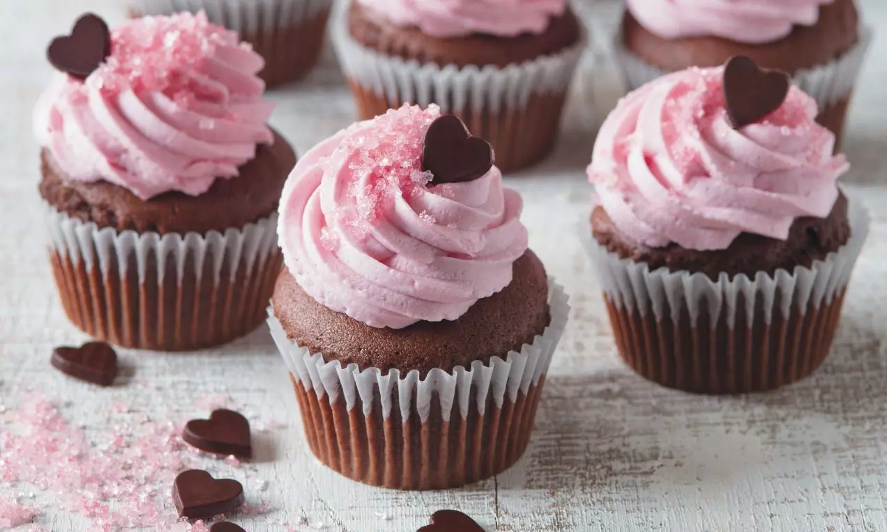 Čokoládové cupcakes v bielych košíčkoch s nadýchaným ružovým krémom, posypané ružovým cukrom a ozdobené tmavými čokoládovými srdiečkami