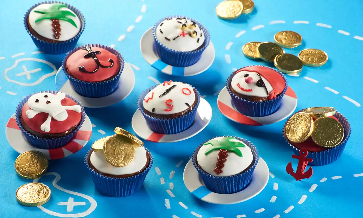Piraten-Muffins mit bunter Fondantdeko wie Schädeln, Ankern und Palmen in blauen Förmchen, umgeben von goldenen Schokomünzen auf blauem Grund