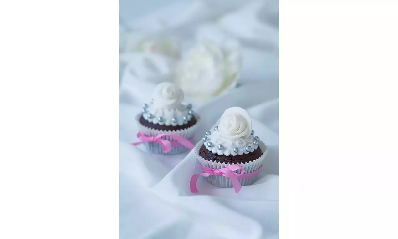 Čokoládové cupcakes s bielou fondánovou ružou, perličkovou ozdobou a ružovou stuhou na jemnom bielom podklade s rozostrenými kvetmi