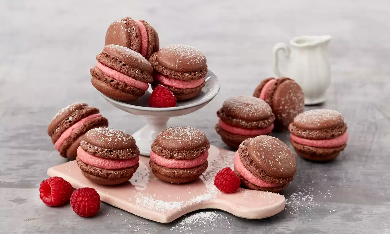 Schokoladenbraune Macarons mit rosafarbener Füllung, leicht mit Puderzucker bestäubt, auf rosa Platte mit frischen Himbeeren angerichtet