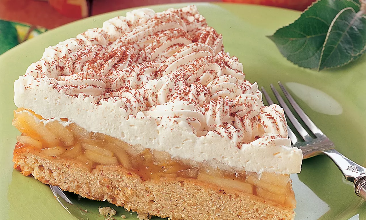 Ein Stück Apfel-Schaumkuchen mit hellbraunem Boden, zarten Apfelscheiben, luftiger weißer Cremeschicht und Kakao auf einem grünen Teller