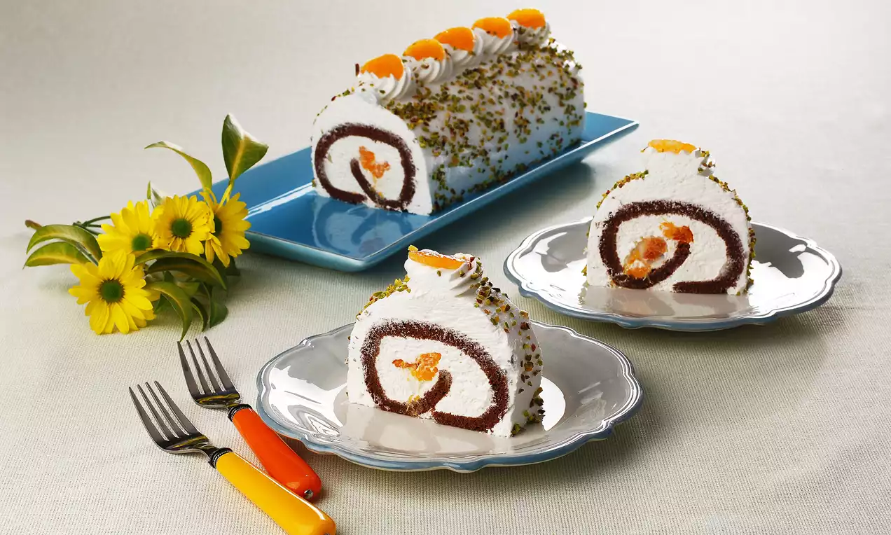 Beyaz kremalı ve yeşil fıstık parçalarıyla kaplı rulo pasta, mandalina dilimleriyle süslenmiş ve iki dilimi porselen tabaklarda sunulmuş