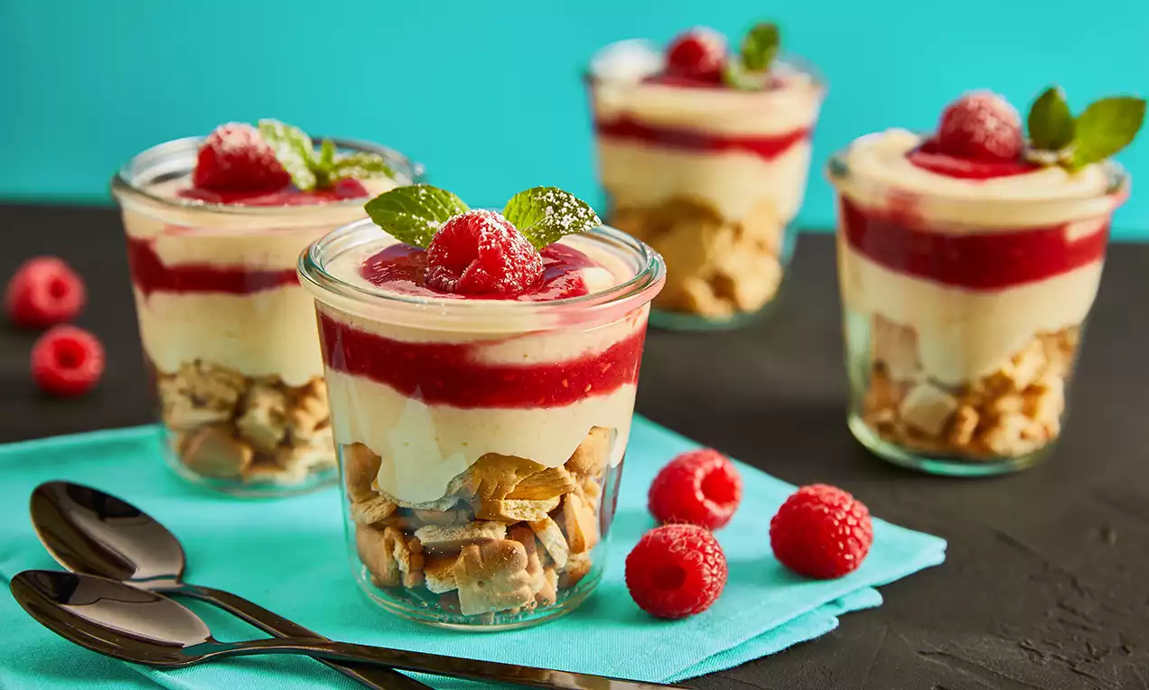 Geschichteter Proteinpudding in einem Glas mit knusprigen Keksstücken, cremiger Vanilleschicht, rotem Himbeerpüree, Beeren und Minze