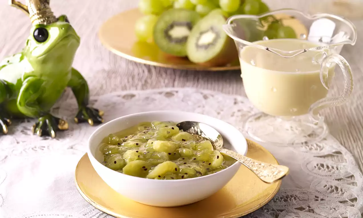 Sofisticiran voćni desert u bijeloj zdjelici, kockice kivija i komadići grožđa s točkicama sjemenki na zlatnom tanjuru, uz vrč sa svijetlom kremom