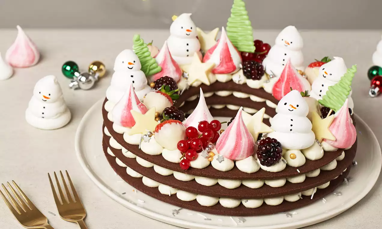 Three stacked layers of rich brown cookie rings with piped white cream, decorated with meringue snowmen, pink swirls, berries, and star shapes on a white plate