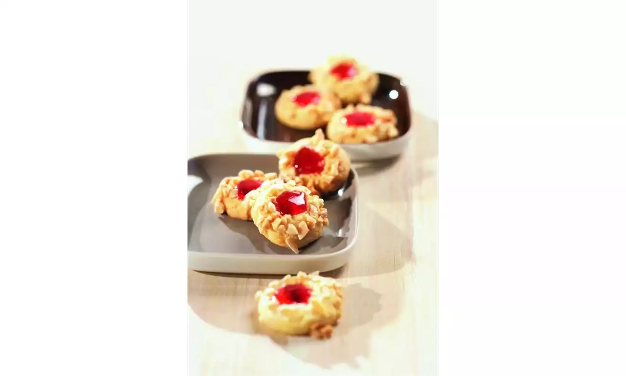 Máslové sušenky světle hnědé barvy s vroubkovaným okrajem, plněné červenou lesklou marmeládou, uspořádané na dvou tmavých talířích