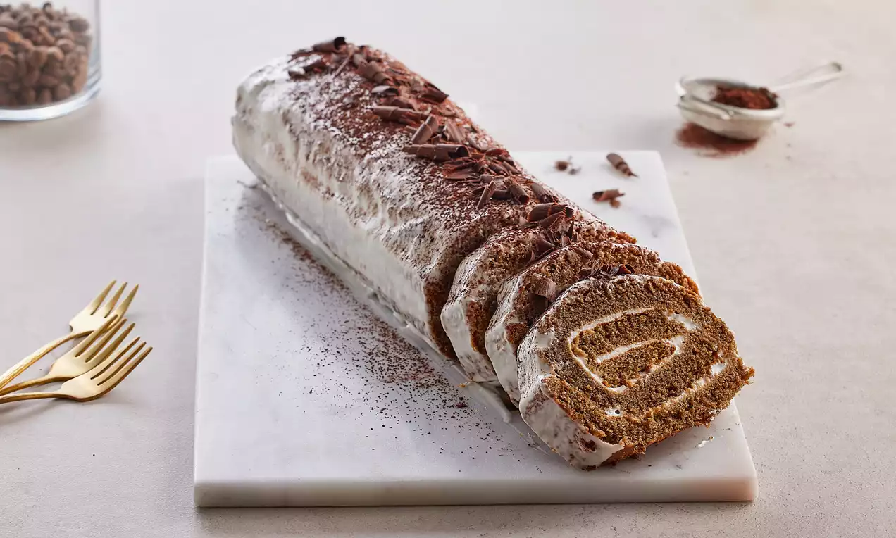 Kahverengi rulo kek dilimleri beyaz krema ile kaplı, üzerine çikolata parçaları serpilmiş ve mermer bir tabakta sunulmuş