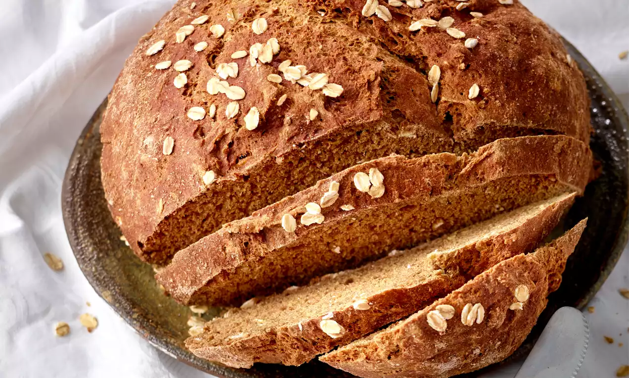 Rund gebackenes, goldbraunes Vollkornbrot mit knuspriger Kruste, Haferflocken bestreut, angeschnitten auf rustikalem Keramikteller