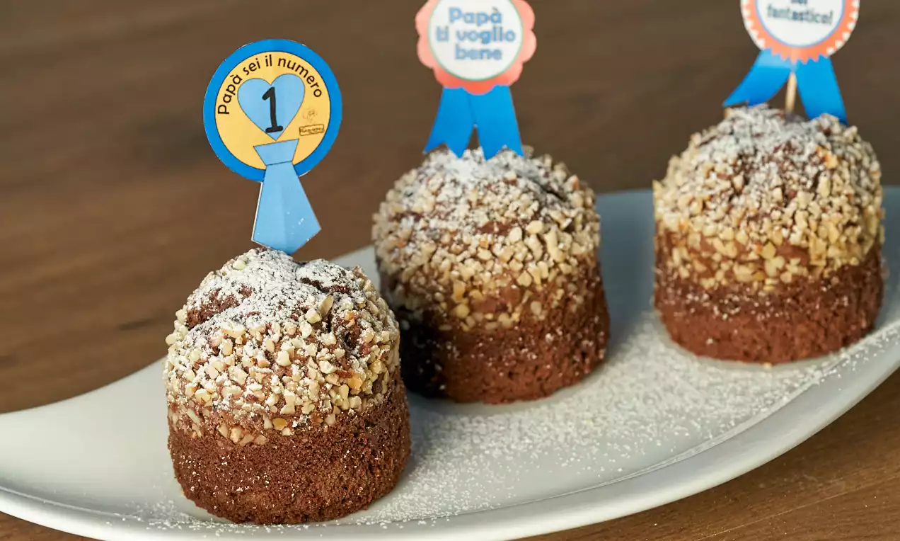 Muffin cilindrici color cioccolato scuro con granella di nocciole dorata, spolverati di zucchero a velo su un piatto bianco