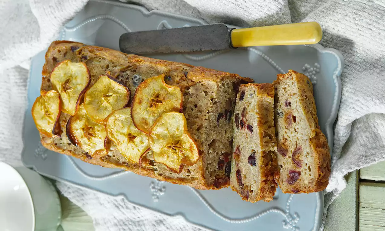 Zlatohnedý jablkový chlebíček s plátkami sušených jabĺk na povrchu, s na krájanie pripraveným nožom na svetlom dekoratívnom podnose