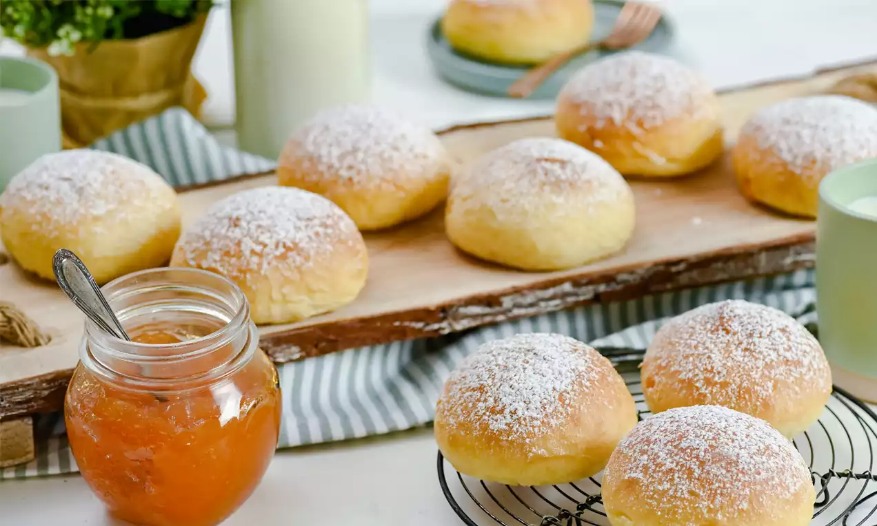 Goldgelbe, rundliche Krapfen mit puderzuckerverzierter Oberfläche auf Holzplatte und Gitter serviert, flankiert von Marmeladenglas und grünen Tassen