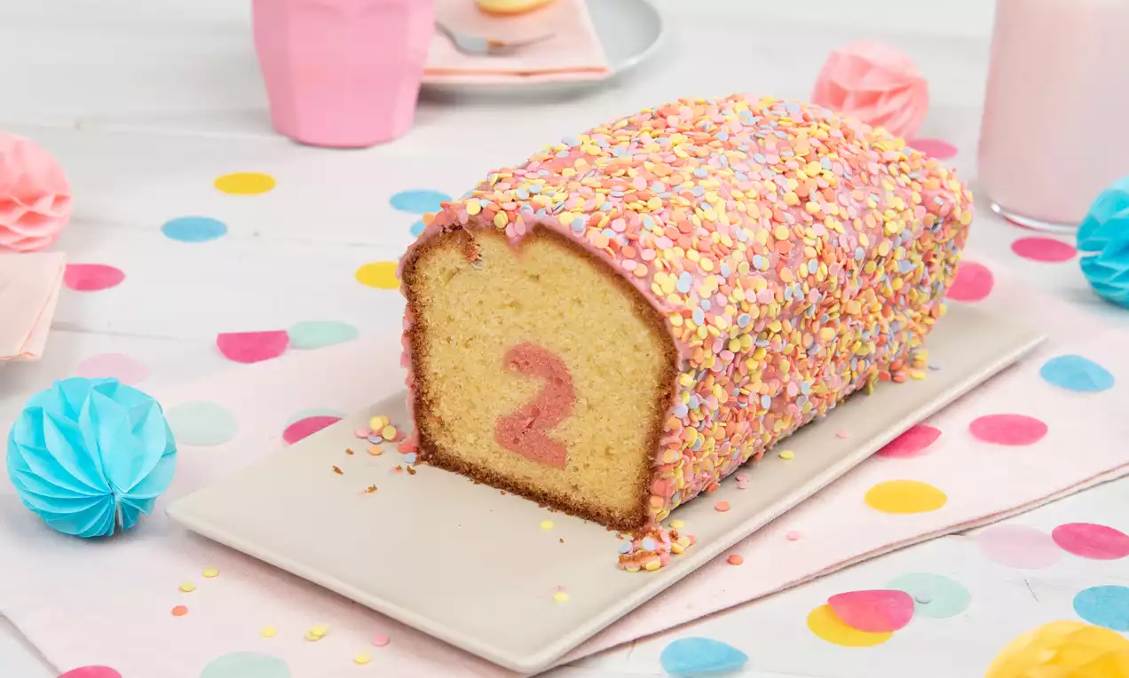 Rosa Überraschungskuchen mit bunter Zuckerstreuselglasur und rosa Zahl im Inneren auf weißer Platte auf dekorativem Tisch mit Pastellfarben