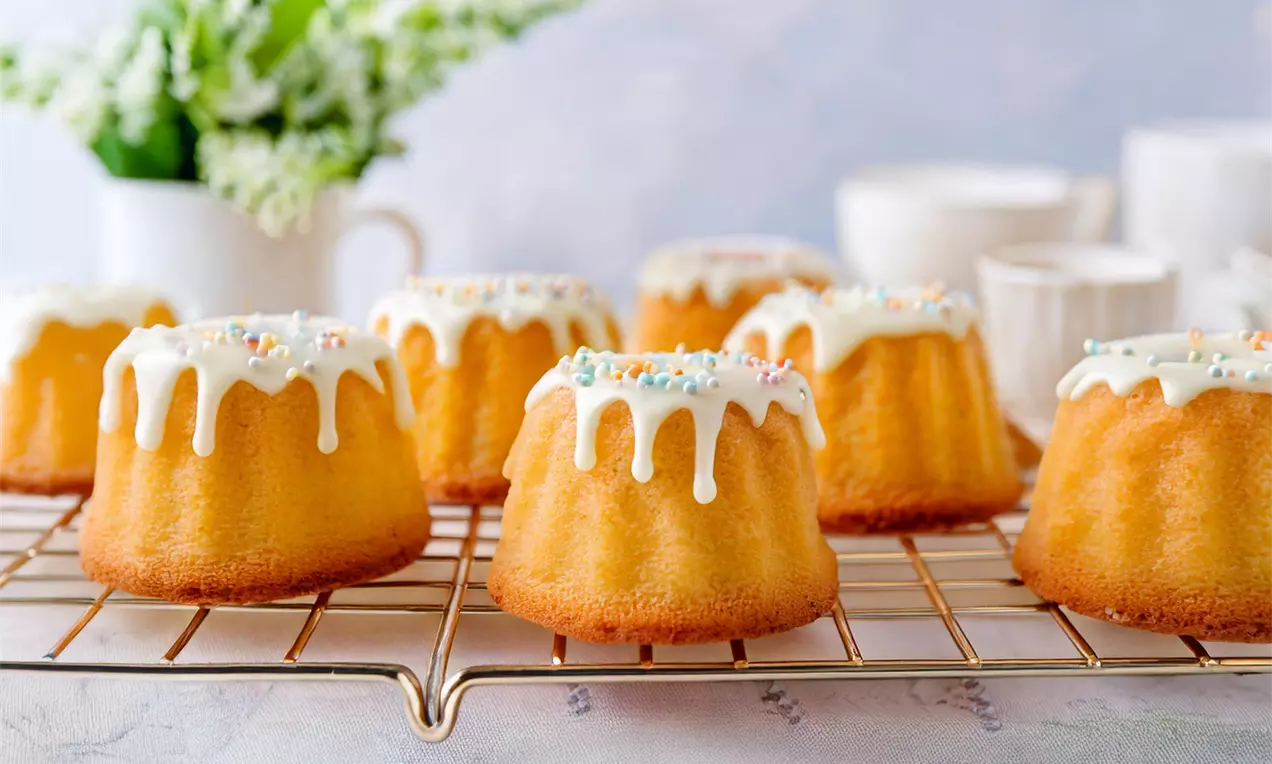 Goldgelbe Mini-Gugelhupfe mit glänzendem weißen Zuckerguss und bunten Streuseln auf einem goldenen Kuchengitter vor pastellfarbenem Hintergrund