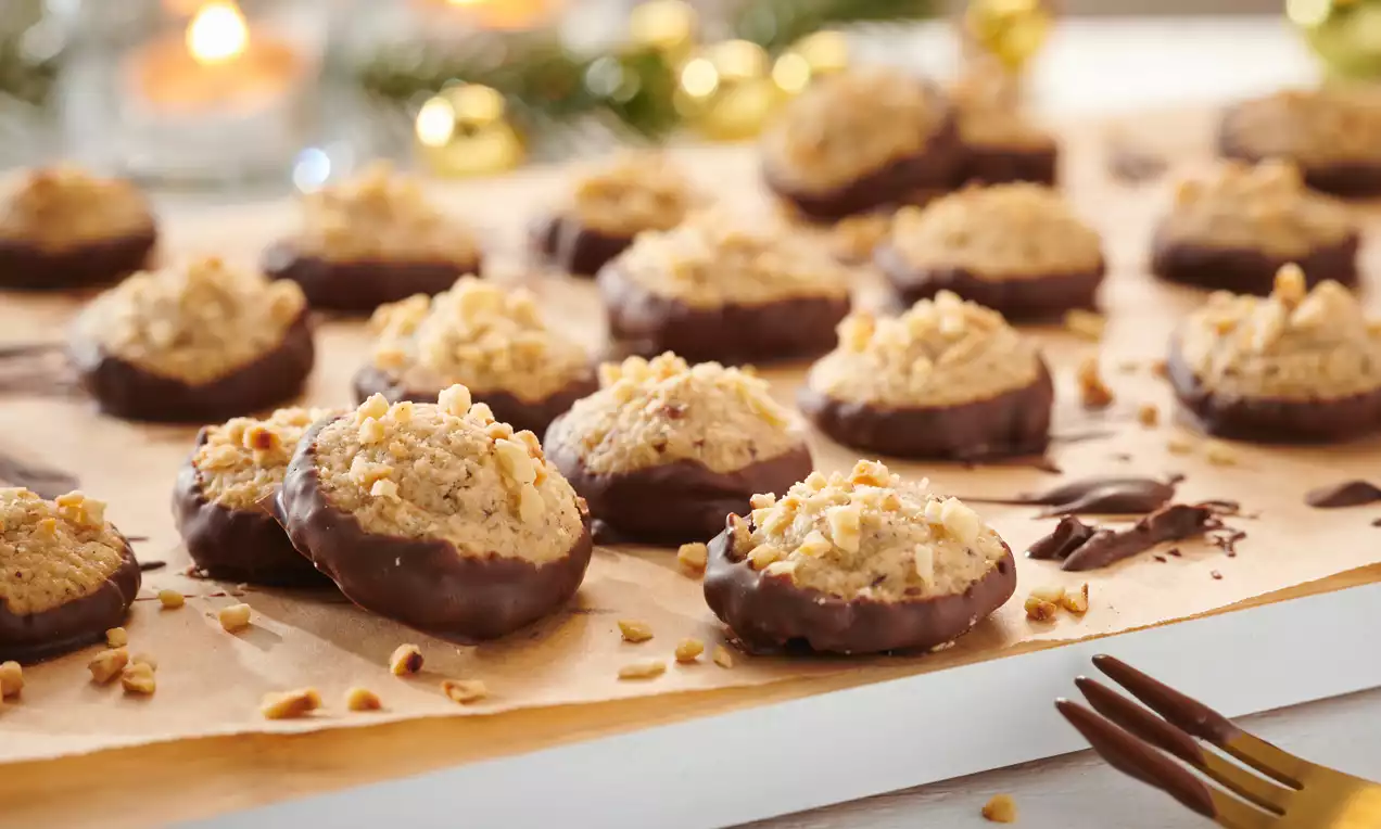 Goldbraune Marzipan-Makronen mit Haselnussstückchen, teils in dunkle Schokolade getaucht, auf Backpapier mit weihnachtlicher Dekoration im Hintergrund