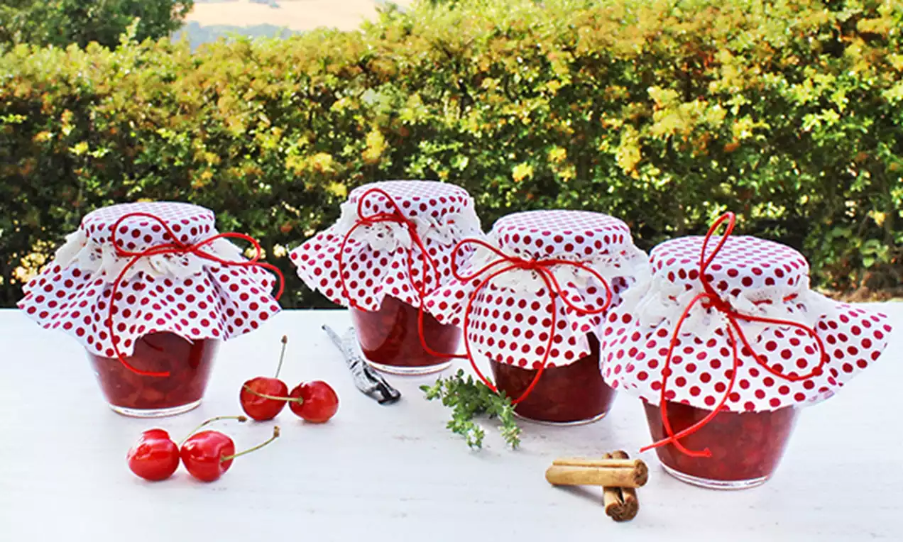 Barattoli trasparenti riempiti con confettura rosso vivo, coperti da teli bianchi a pois rossi annodati con nastri, circondati da ciliegie fresche e cannella