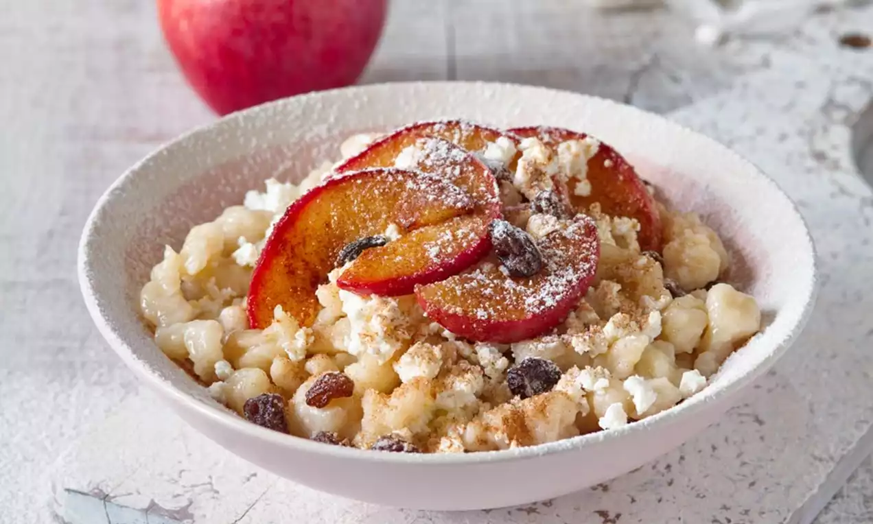 Biely tanier s haluškami krémovej farby, na vrchu plátky opečených jabĺk so škoricovým posypom, hrozienkami a jemnou vrstvou práškového cukru