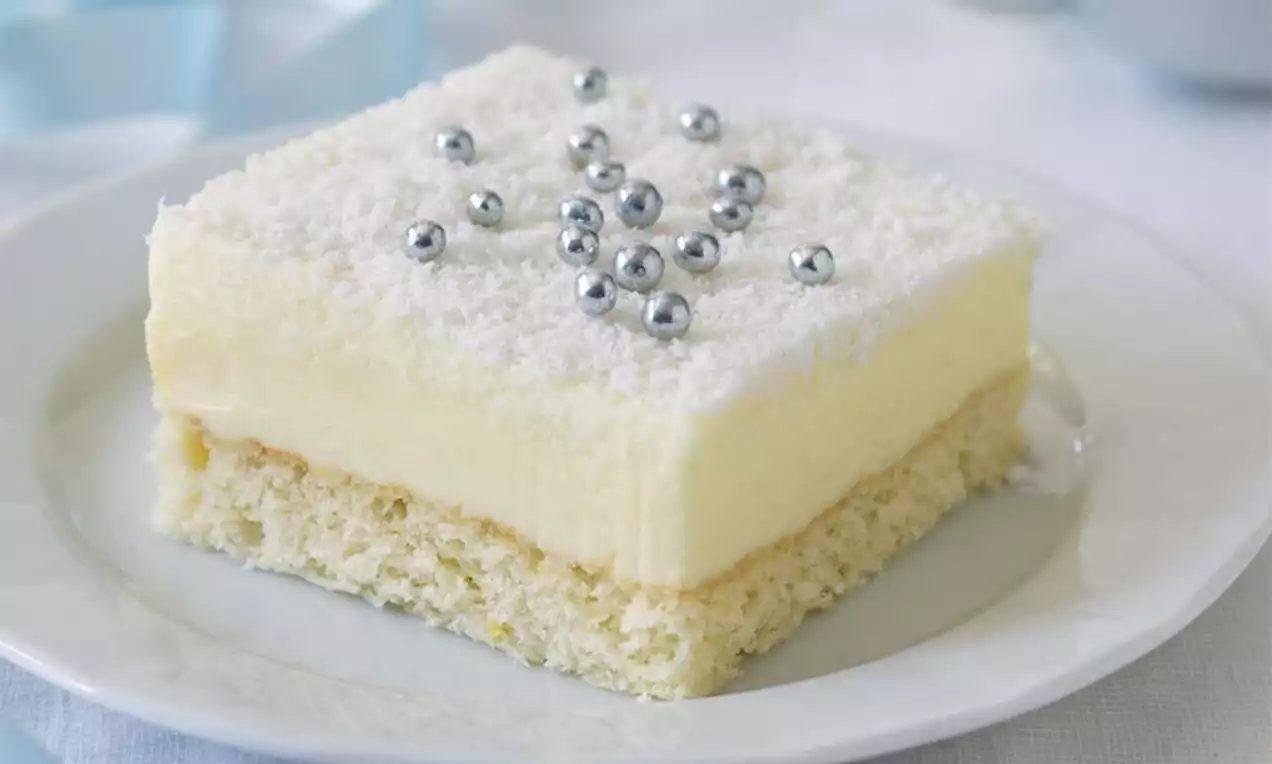 Čtvercový zákusek na bílém talíři s vrstvou světlého krému, posypaný kokosovou moučkou a ozdobený stříbrnými cukrovými kuličkami