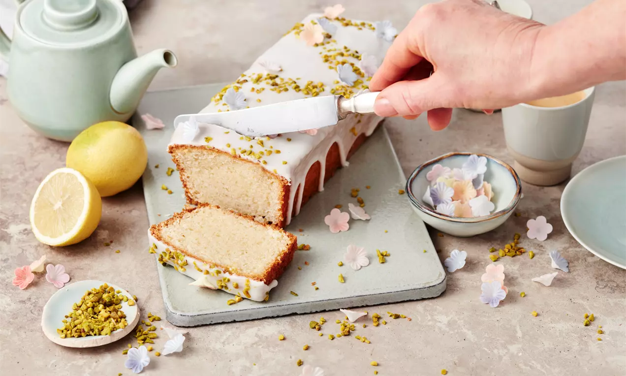 Rechteckiger Zitronenkuchen mit weißem Zuckerguss und gehackten Pistazien, aufgeschnitten auf einer Steinplatte mit Dekoration aus Blüten.