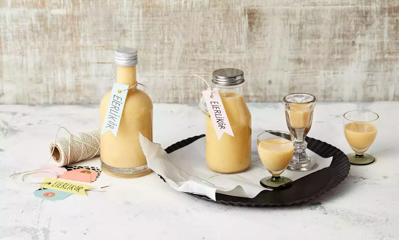 Eierlikör in gläsernen Flaschen mit handbeschrifteten Anhängern neben kleinen Gläsern auf einem schwarzen Tablett mit weißem Papier drapiert