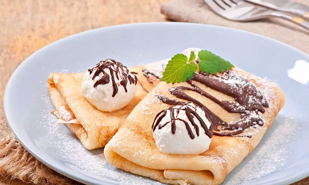 Thin golden crepes folded into rectangles, topped with whipped cream, drizzled chocolate, mint leaves and dusted with icing sugar on a blue plate