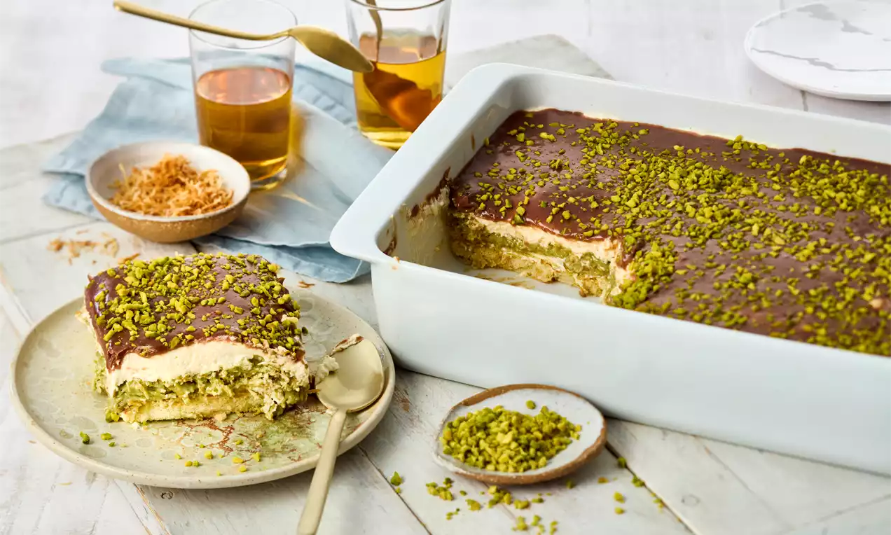 Tiramisu stratificat în vas alb, cu cremă fină, blat însiropat, acoperit cu glazură de ciocolată și presărat din abundență cu fistic