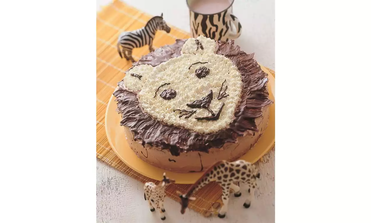 Kruhová torta s hnedým čokoládovým okrajom a bledou kokosovou polevou v podobe levieho tváre na žltom tanieri pri plastových zvieratkách