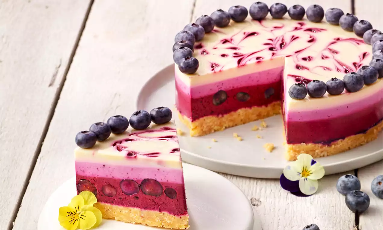 Kleiner Blueberry-Cheesecake mit goldbraunem Keksboden, lilafarbener Beerenfüllung, heller Creme, Heidelbeerenrand und Blumen auf weißem Teller