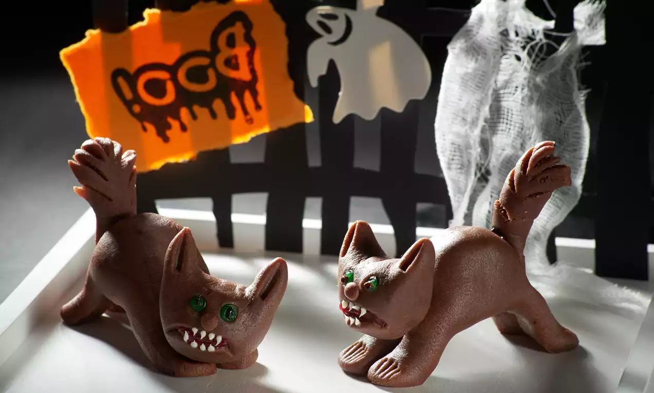 Schokoladenfarbene Katzenfiguren mit grün funkelnden Augen und gezackten Zähnen auf einer weißen Oberfläche vor gruseligem Dekor drapiert