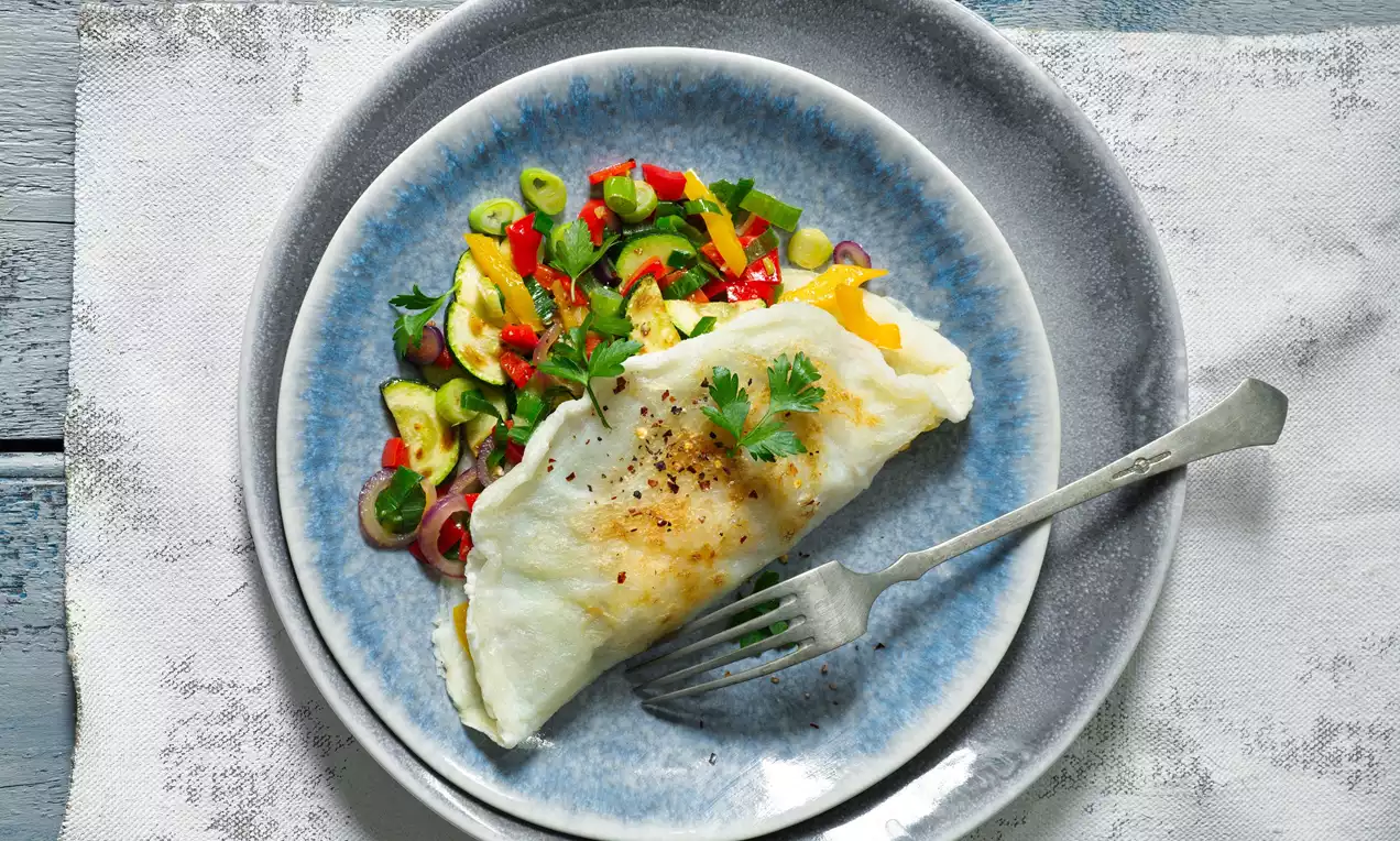 Zlatavá omeleta složená do půlkruhu, posypaná čerstvým pepřem a ozdobená lístky petržele vedle pestrobarevné zeleninové směsi na modrém talíři