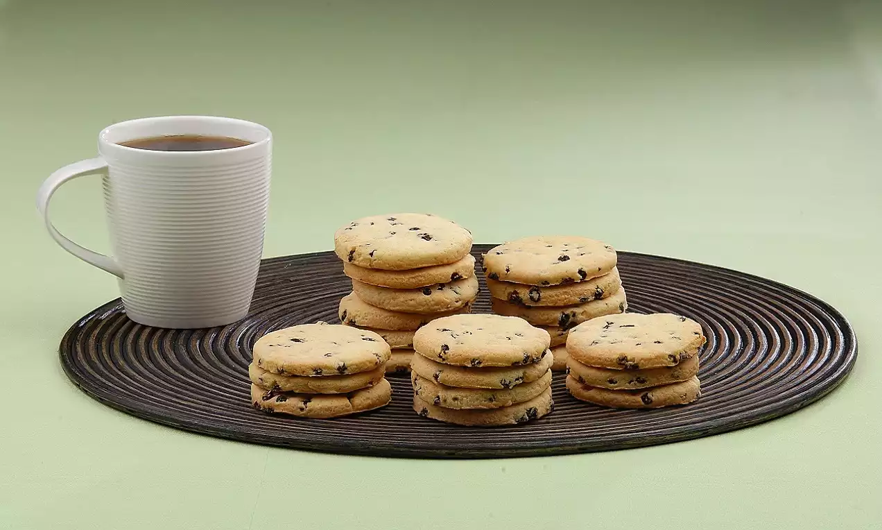 Açık kahverengimsi tonlarda üzüm parçalarıyla dolu yuvarlak kurabiyeler, siyah çizgili tepsi üzerinde bir fincan çay ile sunulmuş