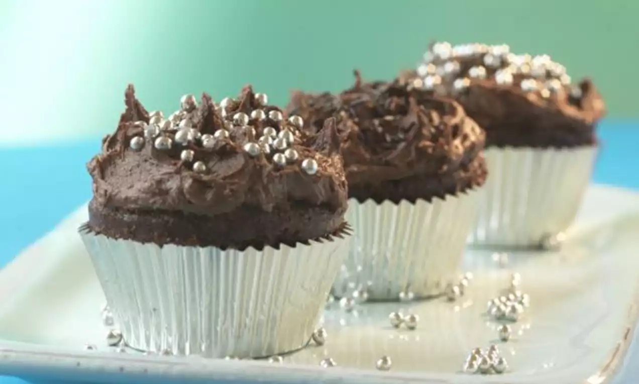 Tre sjokoladecupcakes med mørkebrun krem, sølvfargede sukkerperler og sølvfolieformer på et hvitt fat mot blå og grønne nyanser