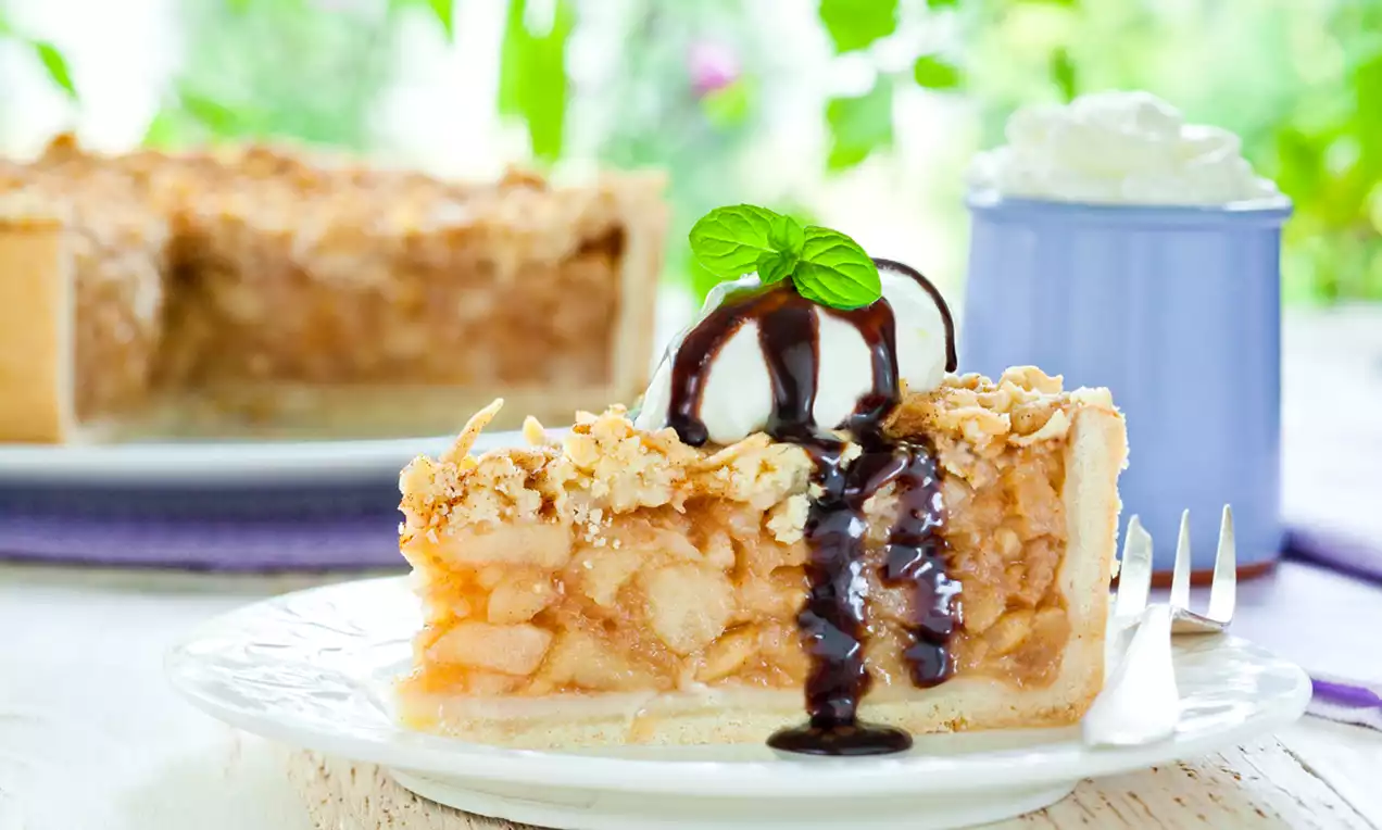 Kawałek szarlotki o złocistym kruchym cieście z kawałkami jabłek, oblany ciemnym sosem czekoladowym, udekorowany listkiem mięty na białym talerzu