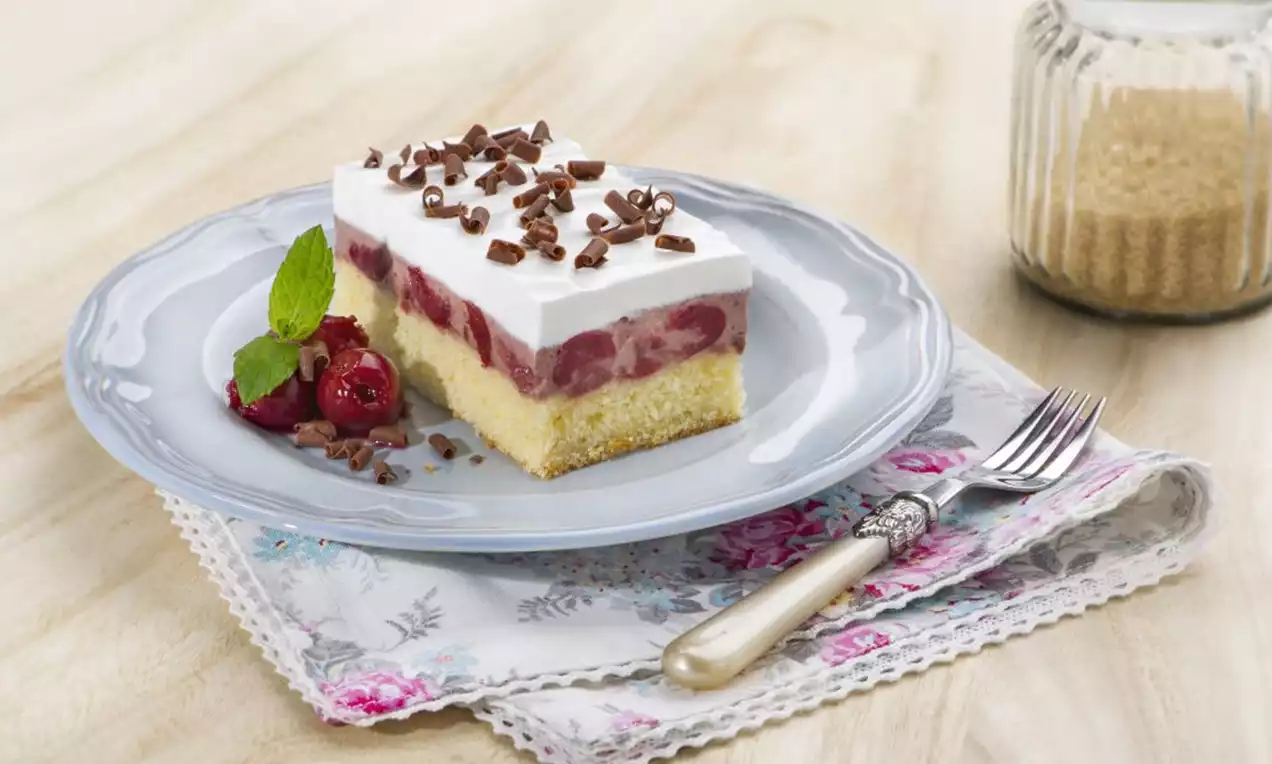 Trojvrstvový zákusok so žltým piškótovým základom, višňovou vrstvou a bielym krémom, ozdobený čokoládovými hoblinami na modrom tanieri