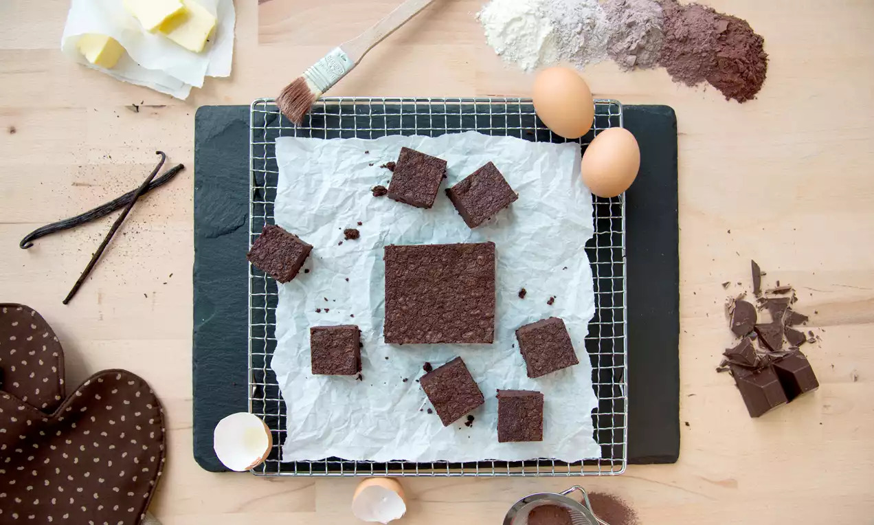 Čokoladni brownies kocke tamnosmeđe boje s glatkom površinom i povremenim mrvicama, raspoređene na bijelom papiru preko rešetke