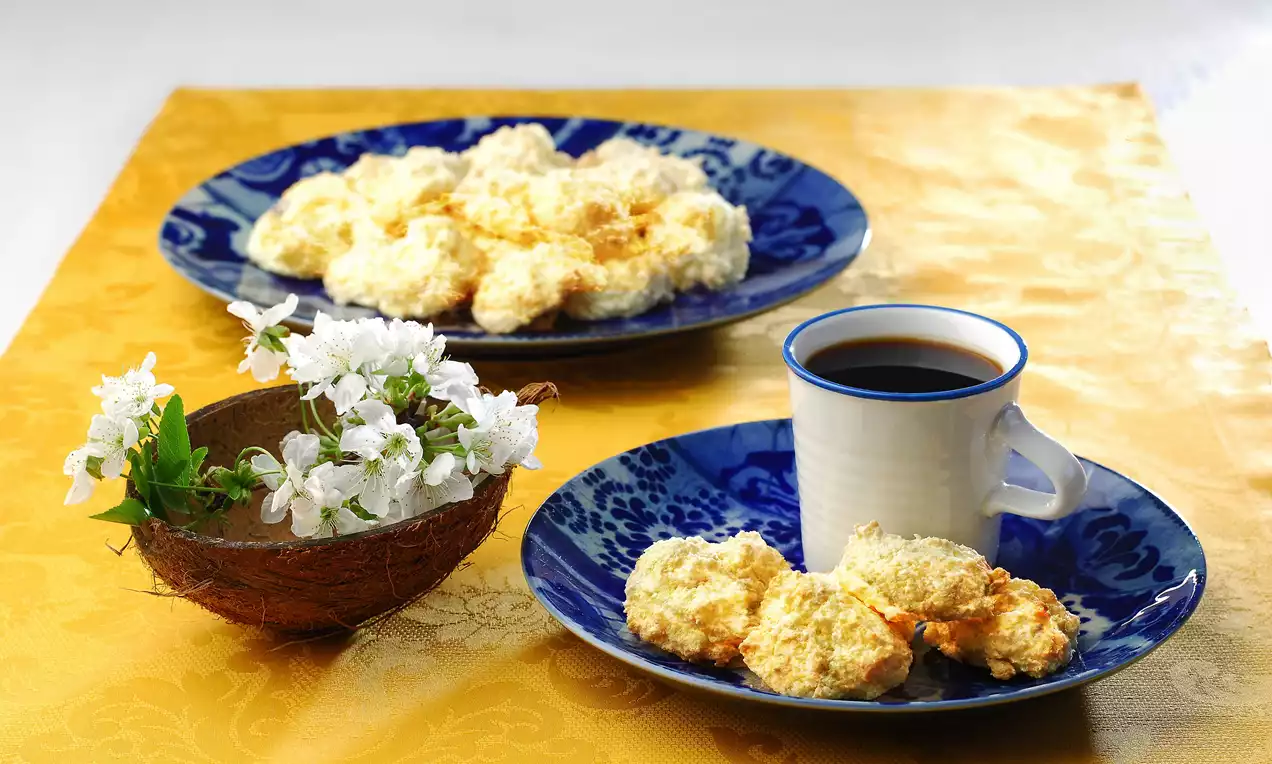 Kokus kurabiyeler altın rengi dokulu, mavi desenli tabakta, kahve fincanı yanında ve hindistan cevizi dekoru eşliğinde sunulmuş