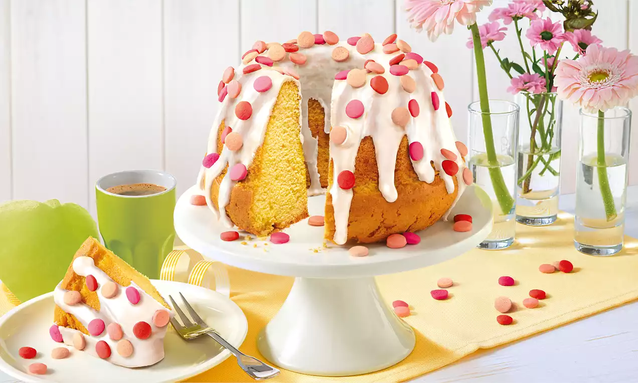 Goldgelber Gugelhupf mit weißer Glasur und rosa sowie roten Schokolinsen auf weißer Tortenplatte, daneben Kuchenstück mit Gabel