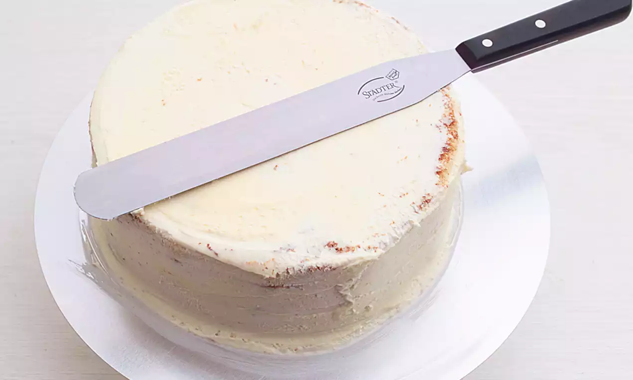 Glatt gestrichene Buttercremeschicht in hellem Beige auf rundem Kuchen, angerichtet auf silberner Platte, mit Metallpalette verziert