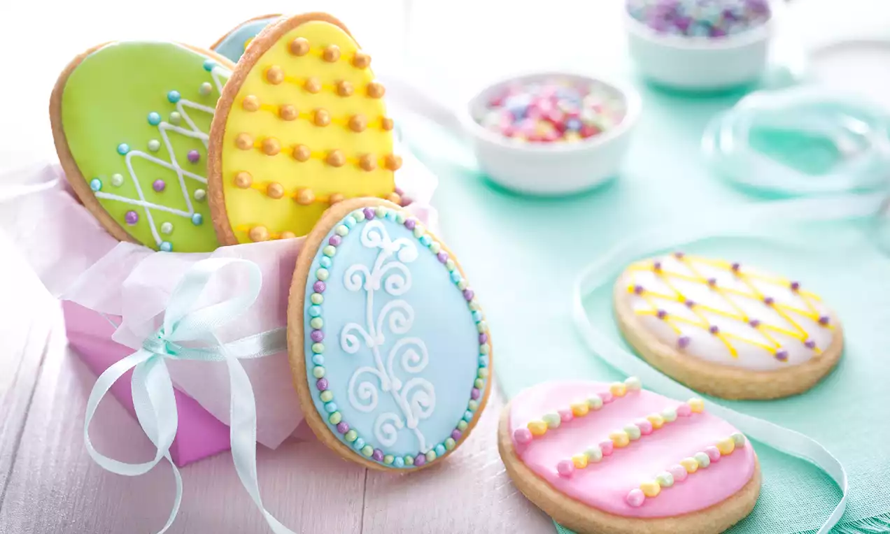 Pääsiäisen munanmuotoisia keksejä pastellinsävyisillä kuorrutteilla, helmikoristeilla ja yksityiskohtaisilla kuvioilla vaaleanpunaisessa rasiassa.