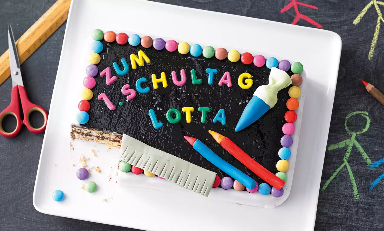 Rechteckige Torte mit schwarzer Glasur, bunten Buchstaben, Zuckerstreuseln und Fondant-Dekoration auf weißer Platte mit Kreidetafel-Hintergrund