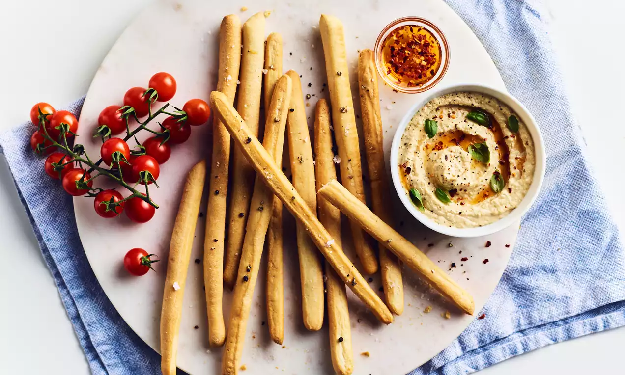 Golden, thin breadsticks speckled with seeds on a white marble plate with creamy hummus, spiced oil and a cluster of red cherry tomatoes