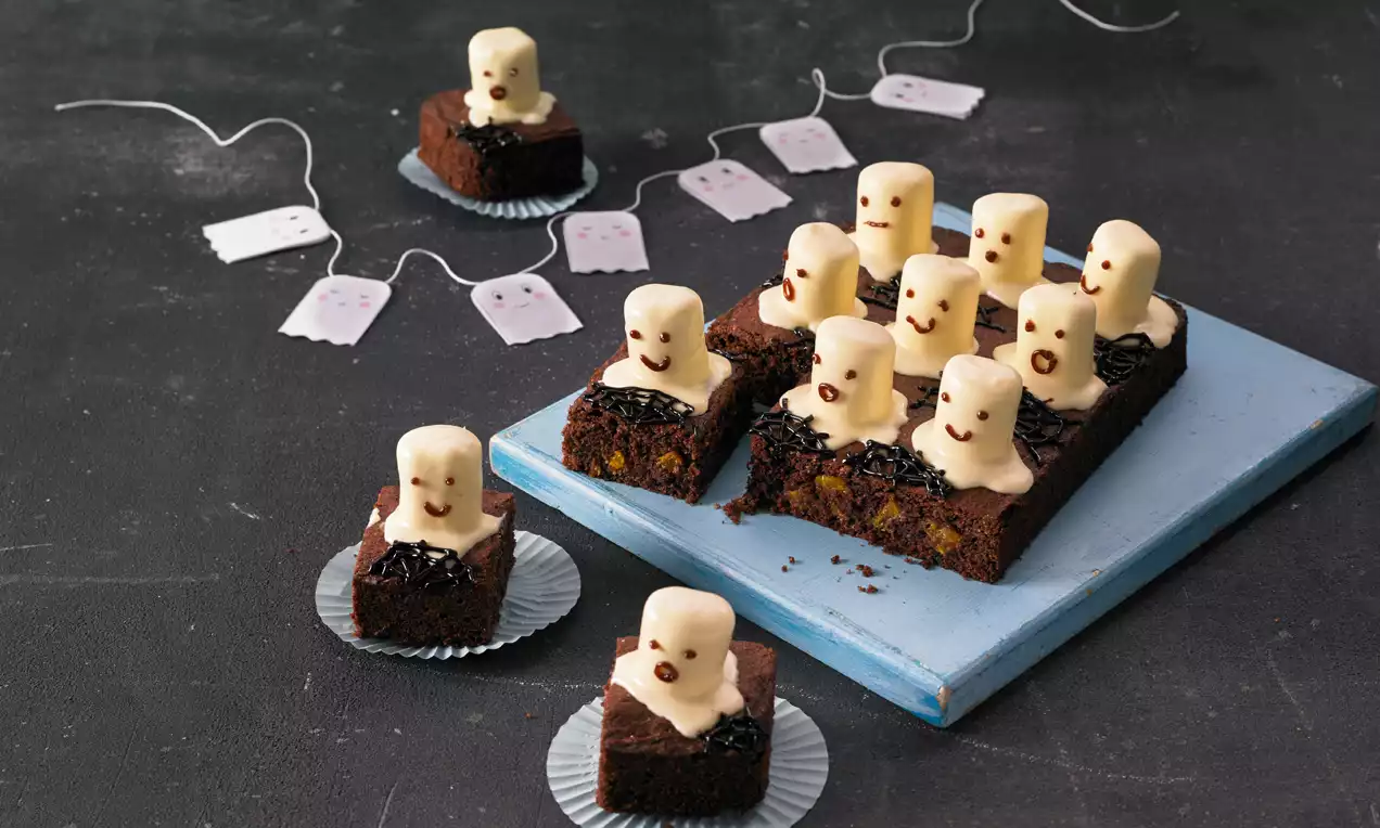Rechteckiger, dunkler Schokokuchen mit weißen Marshmallow-Gespenstern, Schokoglasur und Gesichtsmuster auf blauer Platte und Silberförmchen 