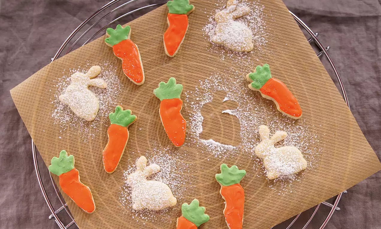 Osterkekse in Hasen- und Karottenformen mit weißem Puderzucker bestäubt und bunter Glasur auf braunem Backpapier, umgeben von einem Gitter