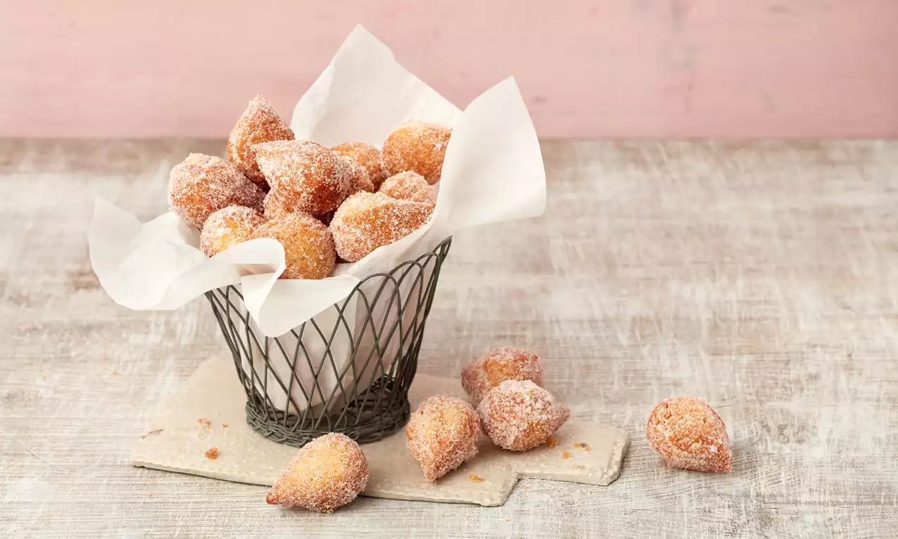 Goldbraune, zuckerbestäubte Muzenmandeln in einem Drahtkorb mit weißem Papier, arrangiert auf hellem Holz mit rosa Hintergrund