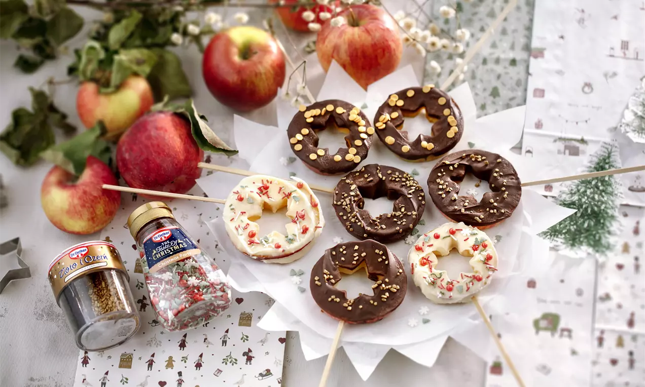 Apaļas ābolu šķēles uz kociņiem, pārklātas ar gludu piena un baltās šokolādes kārtu, rotātas ar zelta un sarkaniem cukura graudiņiem, sakārtotas uz balta šķīvja starp svaigiem āboliem un eglīšu tematiku fona.