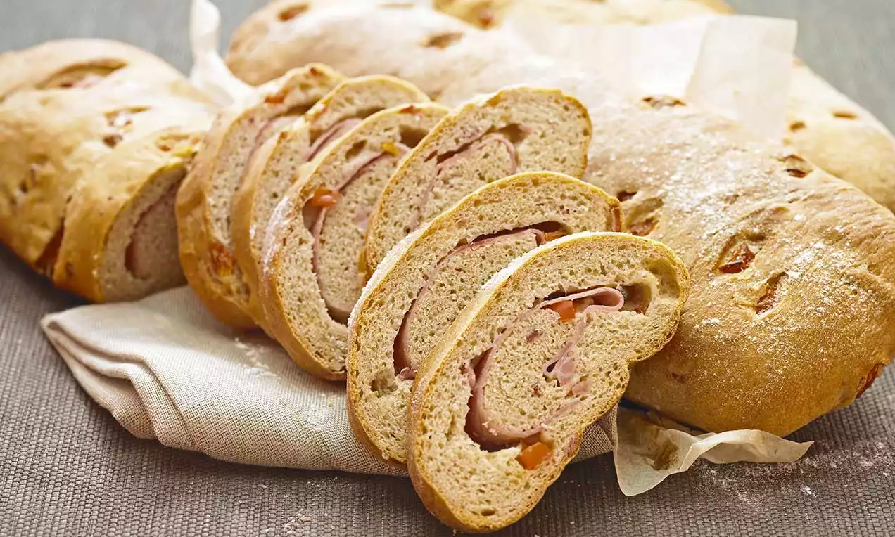 Fette di pane dorato e morbido, arrotolate con prosciutto rosa e guarnizioni, adagiate su un tessuto chiaro e sfondo rustico