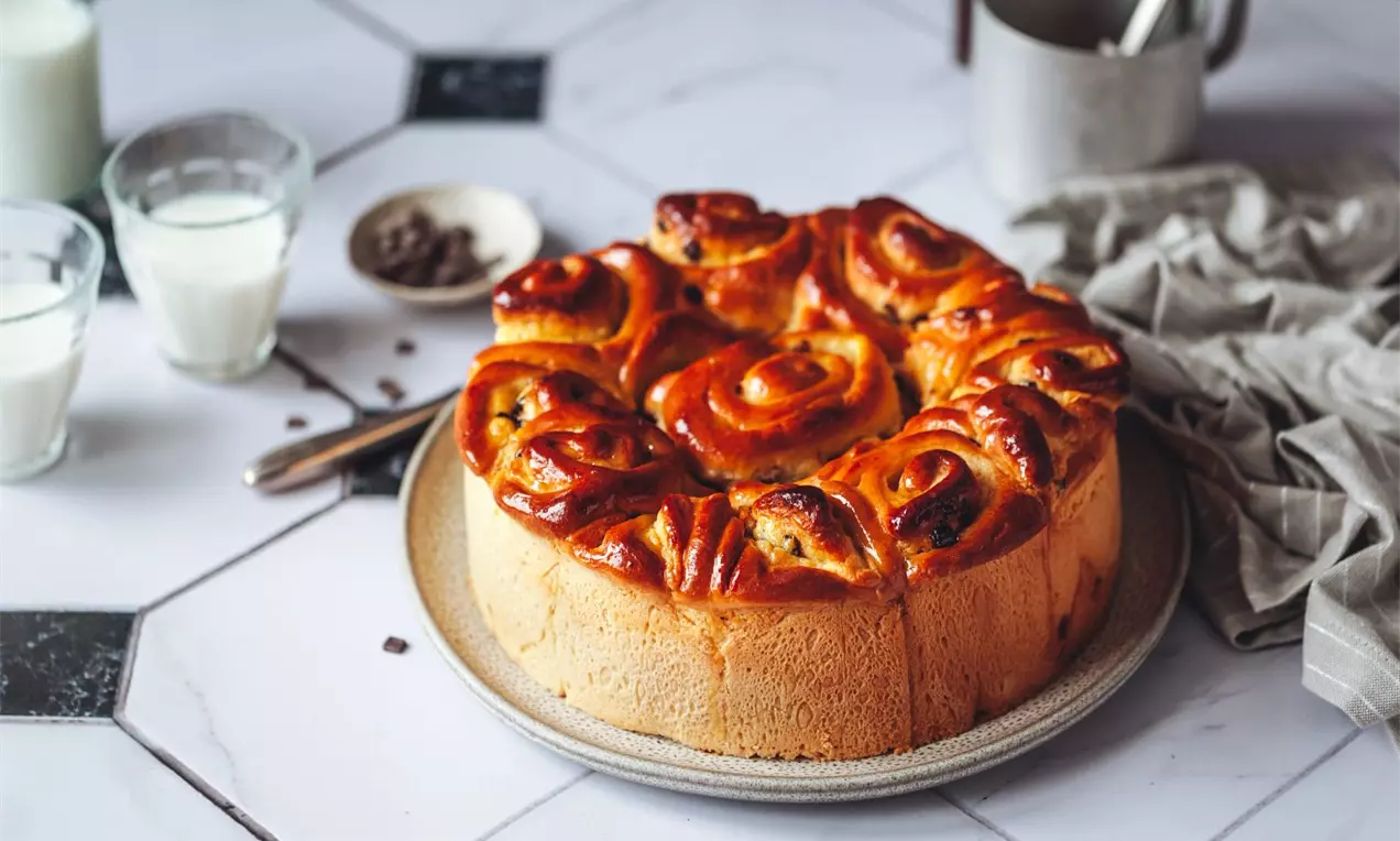 Nadýchaný kulatý koláč s lesklými, dozlatova upečenými záviny plněnými vanilkovým krémem na šedém talíři, na mramorovém stole s mlékem