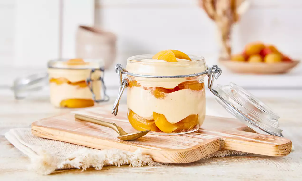 Aprikosen-Dessert mit geschichteter Creme und saftigen Aprikosenstücken in geöffnetem Weckglas auf hellem Holzbrett mit Stoffserviette