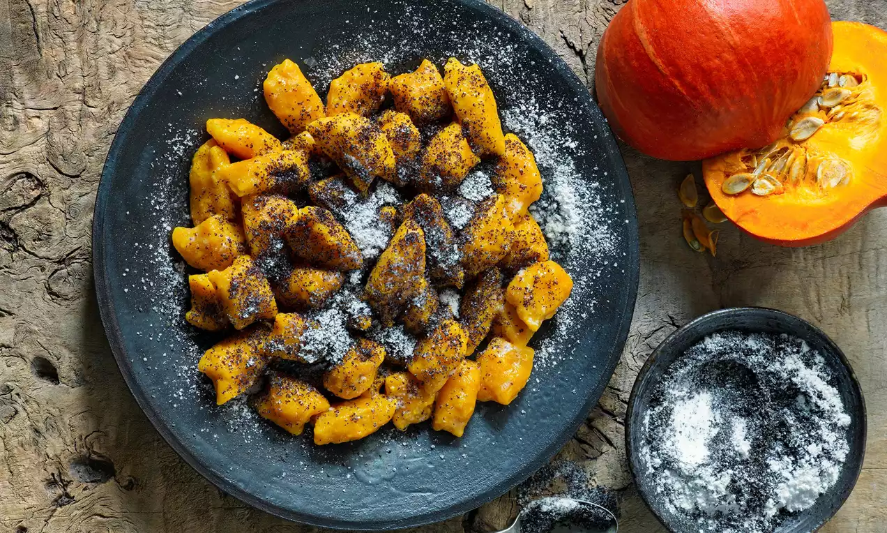 Dýňové noky oranžové barvy, měkké a zaoblené, posypané tmavým mákem a moučkovým cukrem, podávané na černém talíři vedle rozkrojené dýně